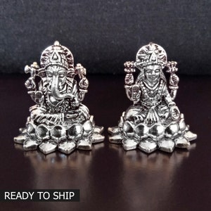 Può includere: Due figurine argentate raffiguranti le divinità indù Ganesha e Lakshmi, sedute su basi di fiori di loto. Le figurine sono dettagliate e intricate, con le divinità raffigurate in pose tradizionali.