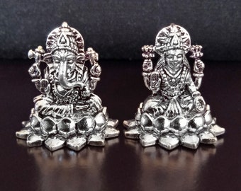 Op de afbeelding: Twee zilverkleurige beeldjes van de hindoe-goden Ganesha en Lakshmi, zittend op lotusbloem-bases. De beeldjes zijn gedetailleerd en ingewikkeld, met de goden afgebeeld in traditionele poses.