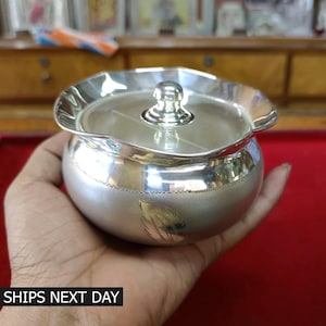 May include: A silver container with a lid and a decorative design. The container is round and has a scalloped edge. The lid has a small knob on top.