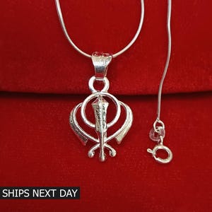 May include: Silver pendant necklace featuring a Khanda symbol, a religious symbol in Sikhism. The necklace has a silver chain.