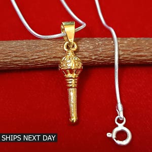 May include: A gold-colored pendant with an ornate design, suspended from a silver chain. The pendant has a detailed, cylindrical shape with intricate patterns. The background is a solid red, with a wooden stick behind the pendant. The text "SHIPS NEXT DAY" is at the bottom.
