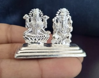 Op de afbeelding: Zilveren beeldje van twee hindoe-goden, Lakshmi en Ganesha, zittend op een zilveren voet. De beeldjes zijn ingewikkeld gedetailleerd en hebben een gepolijste afwerking.