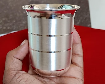 Puede incluir: Un vaso de beber de plata con un acabado cepillado y líneas horizontales alrededor del medio. El vaso se sostiene en una mano.