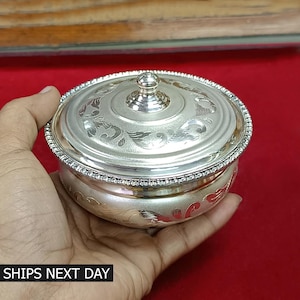 May include: A silver round box with a lid, featuring intricate floral designs engraved on both the lid and the base. The lid has a small knob on top.