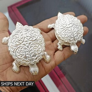 May include: Two silver turtle-shaped boxes with intricate floral designs. The boxes are made of metal and have a shiny finish.