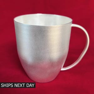 May include: A silver-colored mug with a smooth, metallic finish and a curved handle. The mug features a wide, open top and a rounded base. The text "SHIPS NEXT DAY" is printed in white capital letters at the bottom left of the image.