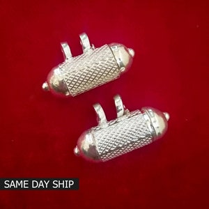 May include: Two silver, cylindrical, decorative charms with a textured surface and a hinged opening.  The charms have a loop at the top for attaching to a necklace or bracelet.