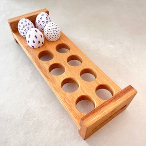Può includere: Un porta uova in legno con 10 scomparti, con quattro uova decorate con sfondi bianchi e motivi geometrici viola. Il porta uova è realizzato in legno marrone chiaro e ha una forma rettangolare.