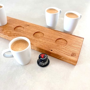 Könnte beinhalten: Ein Holzserviertablett mit vier Espressotassen und einer Kaffeekapsel. Das Tablett besteht aus hellbraunem Holz mit kreisförmigen Vertiefungen für die Tassen. Die weißen Espressotassen sind mit Kaffee gefüllt. Eine schwarze und rote Kaffeekapsel liegt auf der Oberfläche.