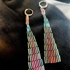May include: A pair of dangle earrings with a gradient design. The earrings feature a series of small, cylindrical beads in shades of teal, pink, and silver. The beads are arranged in a triangular shape, with a silver hoop at the top.