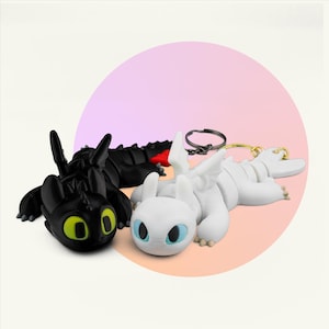 May include: Two dragon figurines, one black with yellow eyes and the other white with blue eyes, are displayed. A black keychain and a gold keychain are also visible. The dragons are lying down, against a pink and peach gradient background.