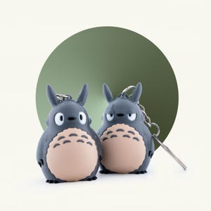 May include: Two gray Totoro keychains with tan bellies and black accents. Each keychain has a silver chain and a silver key ring. The background is a light cream color with a green circle behind the keychains.