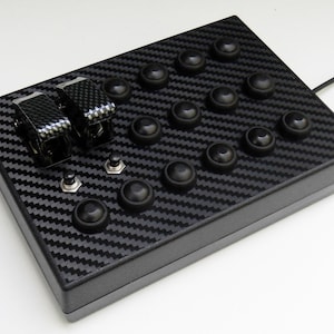 May include: A black control panel with a carbon fibre pattern. It features two toggle switches, several black buttons, and two silver switches. A black cable extends from the side. The device is likely used for gaming or simulation.