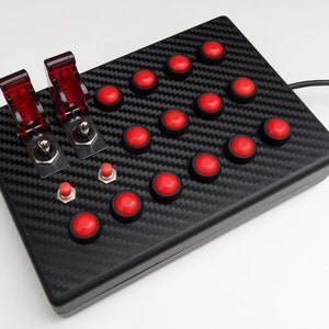 May include: A black gaming controller with a carbon fibre pattern. It has 18 red buttons and two red toggle switches.