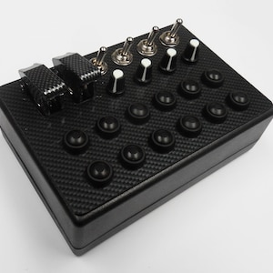 May include: A black control panel with a carbon fibre finish. It has 16 black buttons, 5 toggle switches, and 4 knobs with white caps.