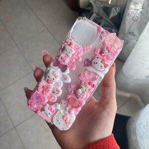 Può includere: Una custodia per telefono rosa e bianca decorata con personaggi di Hello Kitty, fiocchi e cuori. La custodia ha uno sfondo trasparente ed è adornata con vari abbellimenti, creando un effetto 3D. La custodia è progettata per uno smartphone.