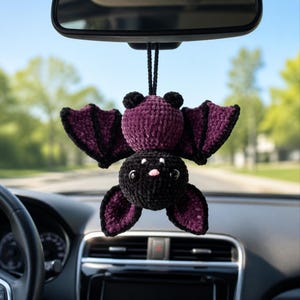 Pode incluir: Um morcego de crochê para pendurar com cabeça preta, olhos brancos e nariz rosa, corpo de pelúcia roxo escuro e asas roxas com bordas pretas. O morcego está suspenso por um cordão preto e apresenta uma textura macia e aveludada.