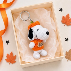 May include: A crochet keychain featuring a white dog with black ears, wearing an orange sweater and an orange pumpkin-shaped hat. The sweater has a small wooden tag with a witch hat icon and the text Happy Halloween printed in black ink.