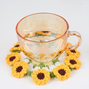 Könnte beinhalten: Eine klare Teetasse aus Glas steht auf einem handgefertigten Häkeluntersetzer. Der Untersetzer hat ein Sonnenblumenmuster mit gelben Blütenblättern, braunen Zentren und grünen Blättern. Die Teetasse hat einen goldenen Farbton.