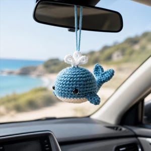 May include: A small, blue, crocheted whale ornament hanging from a light blue string. The whale features a white crocheted water spout on its head, a cream-colored underbelly, and two black, round, glossy eyes. It is designed to hang from a rearview mirror.
