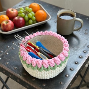May include: A handmade crochet bowl with pink and green floral trim, filled with crochet hooks. The bowl sits on a metal table next to a tray of fruit and a mug of coffee. The bowl is white with pink and green trim.