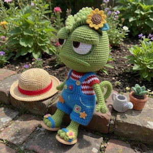 Puede incluir: Muñeca de camaleón de ganchillo verde con un girasol como adorno para la cabeza, con un peto azul y sandalias. Un sombrero de paja con una banda roja está cerca. También hay una pequeña regadera y una planta en maceta.