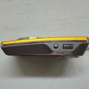 May include: A small, gray and gold digital camera with a black strap. The camera has a power button and a zoom button on the side.