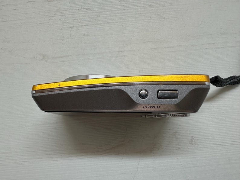 May include: A small, gray and gold digital camera with a black strap. The camera has a power button and a zoom button on the side.