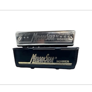 May include: A silver harmonica with the text "Melody Star" and "Hohner" is displayed above a black case with the same text in gold. The harmonica has decorative stars and a shiny finish. The case is rectangular.