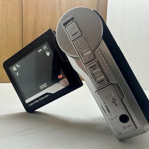 Op de afbeelding: Een zilveren en zwarte digitale videocamera met een klein LCD-scherm waarop "0002" en de tekst "Digital Video Camcorder" wordt weergegeven. De videocamera heeft knoppen voor video, menu, OK, modus, SD/MMC en USB.