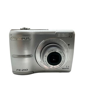 May include: Silver Olympus FE-210 digital camera. The camera has a lens with the text "OLYMPUS LENS" and "OPTICAL ZOOM 3X". The camera also has the text "7.1 MEGAPIXEL".