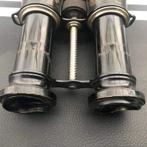 K&ouml;nnte beinhalten: Vintage-Fernglas in Schwarz mit einem silberfarbenen Mittelgelenk und graviertem Text auf den Rohren, der "REG. NO. 151974" und "REG. NO. 151974" lautet.