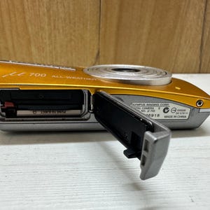 May include: A gold and silver Olympus digital camera with the model number 700 and the text "ALL WEATHER" printed on the front. The camera has a lens cover that is open and a black memory card slot.