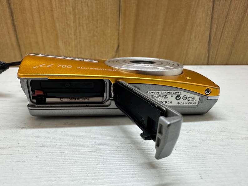 May include: A gold and silver Olympus digital camera with the model number 700 and the text "ALL WEATHER" printed on the front. The camera has a lens cover that is open and a black memory card slot.