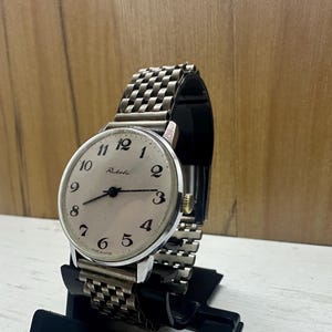 Puede incluir: Un reloj de pulsera plateado con una esfera blanca y números negros. El reloj tiene una correa de metal plateado y dice "Raketa" en la esfera.