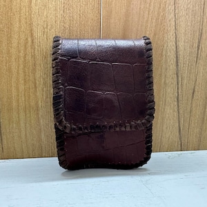 Puede incluir: Un estuche de cigarrillos de cuero marrón con un patrón de cocodrilo y un borde cosido.