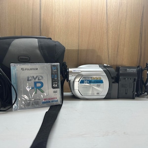 Puede incluir: Una videocámara plateada y negra con un disco DVD-R, una funda de transporte negra y un cargador negro con un cable de alimentación.