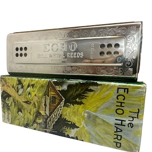 Könnte beinhalten: Eine silberfarbene Metall-Mundharmonika mit eingravierten Blumenmustern und dem Text ECHO BELL METAL REEDS. Sie liegt auf einer rechteckigen Pappschachtel mit einer gemalten Landschaft eines Hauses in den Bergen und dem Text The Echo Harp.
