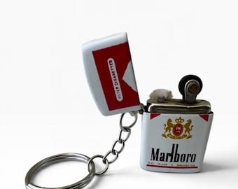 Könnte beinhalten: Ein Metall-Schlüsselanhänger mit einem Miniatur-Feuerzeug in Form einer Marlboro-Zigarettenschachtel. Das weiß-rote Gehäuse zeigt das Marlboro-Logo, den Text Filter Cigarettes und ein goldenes Wappen. Der Feuerzeugmechanismus ist sichtbar.