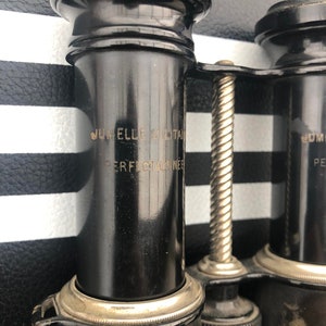 K&ouml;nnte beinhalten: Schwarzes Fernglas mit einem silbernen Scharnier und einem silbernen Einstellknopf. Das Fernglas tr&auml;gt die Gravur "Jumelle Militaire Perfectionnee" auf der Seite.