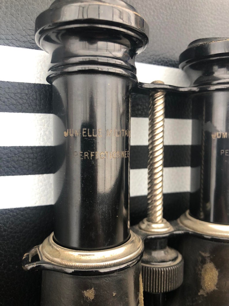 K&ouml;nnte beinhalten: Schwarzes Fernglas mit einem silbernen Scharnier und einem silbernen Einstellknopf. Das Fernglas tr&auml;gt die Gravur "Jumelle Militaire Perfectionnee" auf der Seite.