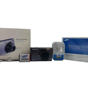 May include: A Samsung digital camera kit, including a black camera, battery, charger, and manuals. The box reads "Never miss another smiling face". The camera is black with a silver lens.