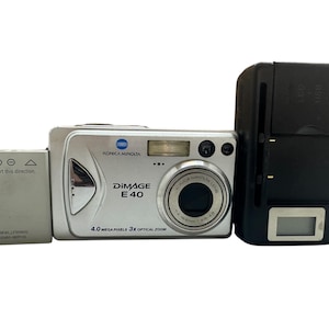 May include: A silver Konica Minolta DiMAGE E40 digital camera with a 4.0-megapixel resolution and 3x optical zoom. Includes a battery and a black charger. The battery has the text "Insert this direction."