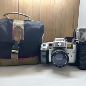 May include: A silver Sony FMD system camera with a flash and a black camera bag. The camera bag is black with a brown and tan base and a brown handle. The camera has a black lens and the text "FMD system" is visible.