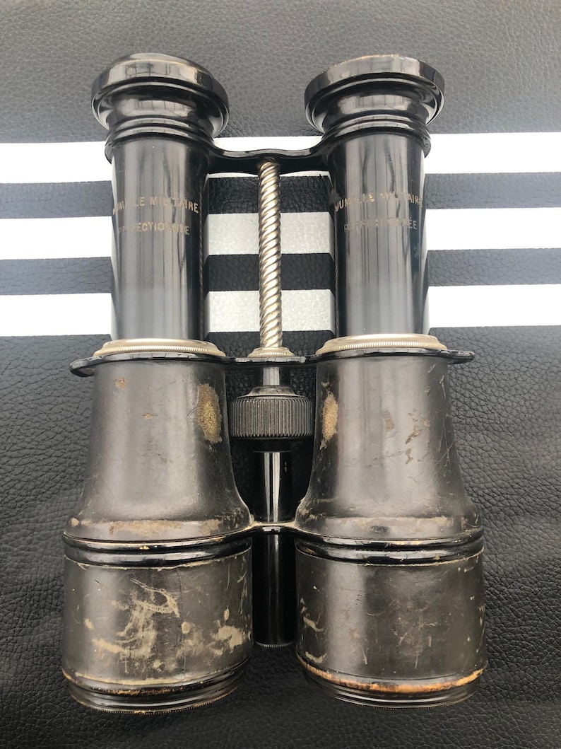 K&ouml;nnte beinhalten: Schwarze Leder-Ferngl&auml;ser mit einem silbernen Mittelgelenk und der Aufschrift "Jumelle Le Militaire Brevetee" auf jeder Seite.