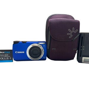 May include: A blue Canon digital camera with a 5x optical zoom lens, a 2GB blue memory card, a black Bluemax battery, a purple zippered camera case with a white logo, and a black battery charger with a small digital display screen.
