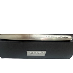 May include: A brushed silver fountain pen with a metallic finish resting on a black rectangular box. The pen features an engraved inscription that reads "Kwei Namen, ein Pilgerhaus" and the box displays a silver Lamy logo plate on the front.