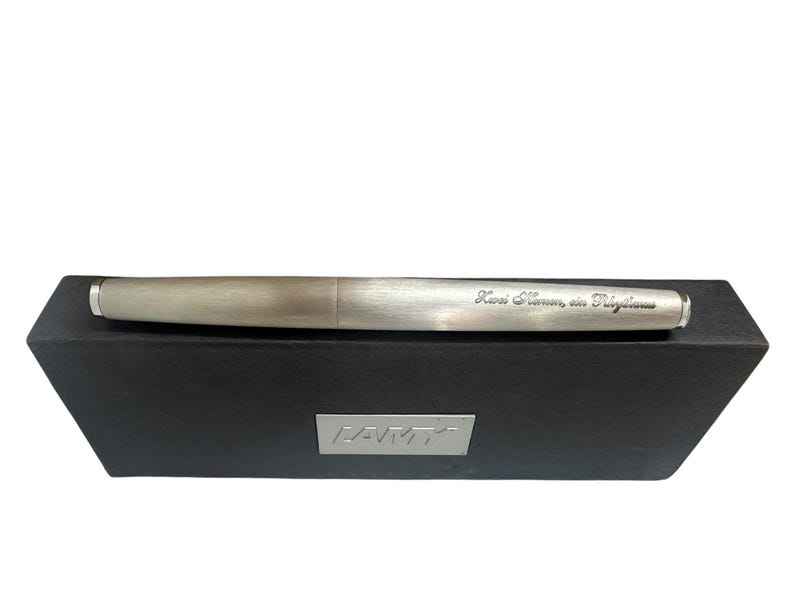 May include: A brushed silver fountain pen with a metallic finish resting on a black rectangular box. The pen features an engraved inscription that reads "Kwei Namen, ein Pilgerhaus" and the box displays a silver Lamy logo plate on the front.