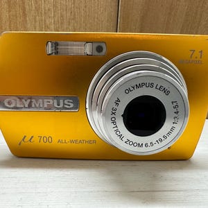 May include: A gold Olympus &micro; 700 digital camera with a 7.1 megapixel lens. The camera has a 3x optical zoom lens with a range of 6.5-19.5mm and a 1:3.4-4.5 aperture. The camera is labeled "All-Weather".