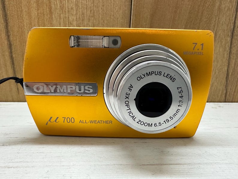 May include: A gold Olympus &micro; 700 digital camera with a 7.1 megapixel lens. The camera has a 3x optical zoom lens with a range of 6.5-19.5mm and a 1:3.4-4.5 aperture. The camera is labeled "All-Weather".